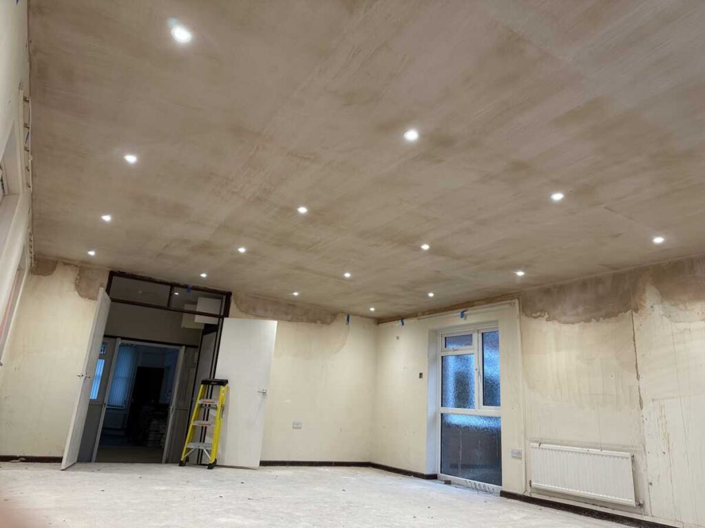 A large, unfinished room with bare plastered walls and ceiling, featuring multiple recessed ceiling lights installed by a Gloucestershire electrician near me, a white door, a window, a radiator, and a yellow step ladder near the far doorway.
