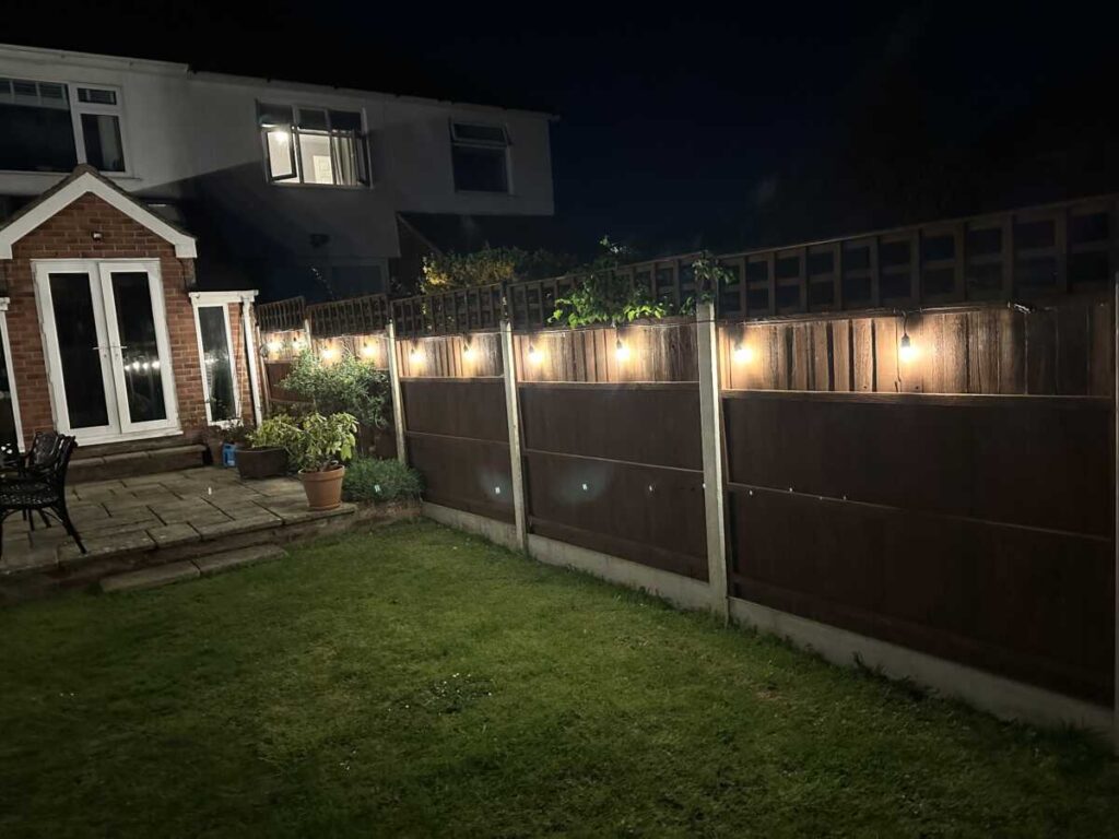 A garden at night with fairy lights, professionally installed by a Gloucestershire electrician near me, illuminating a wooden fence, lawn, patio with chairs, potted plants, and a brick house in the background.