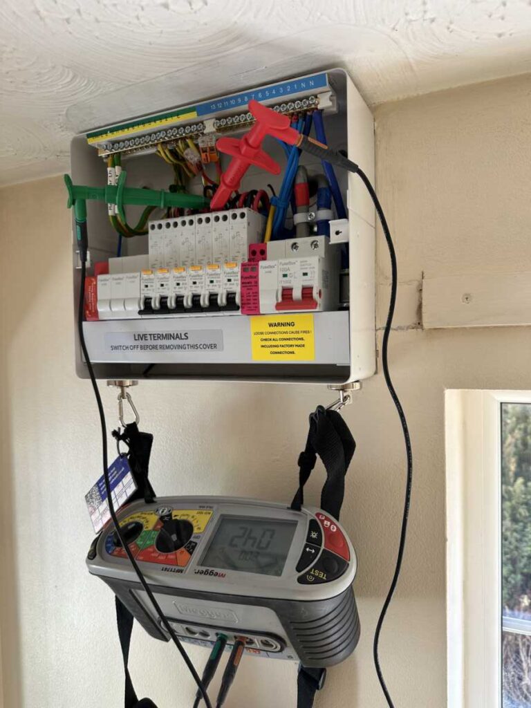 An open electrical consumer unit with coloured wires and circuit breakers exposed. A digital multimeter hangs below, displaying a reading of 246.0. The unit is mounted on a beige wall near a window.